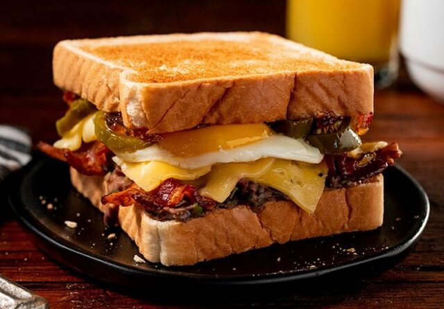 A substantial breakfast sandwich on golden Texas toast with a black bean spread, bacon, aged Gouda, properly scrambled eggs, and jalapeno.